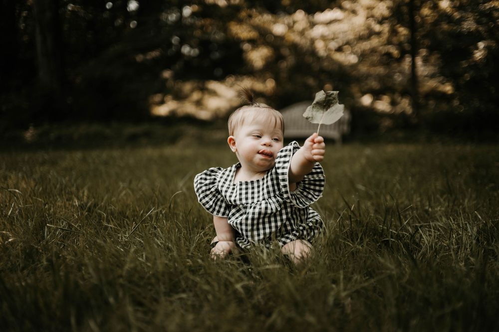 A happy baby girl in a gingham dress sits in green grass, holding a small flower outdoors.