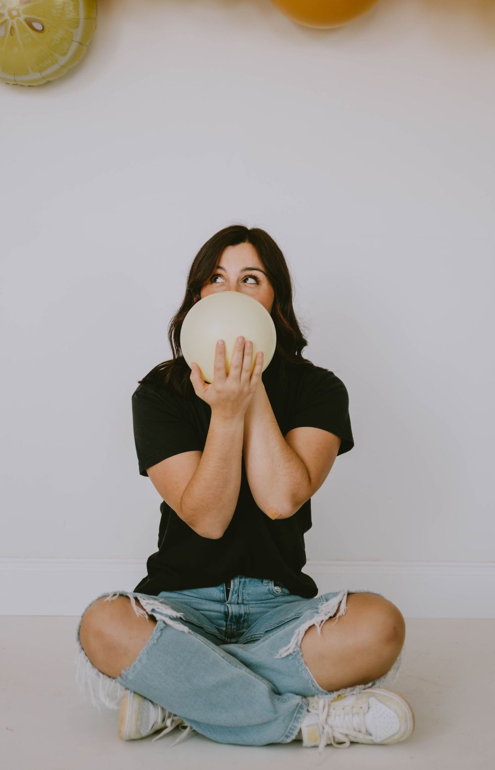 Woman in black tee and ripped jeans sits cross-legged holding a yellow balloon near her face, looking up.