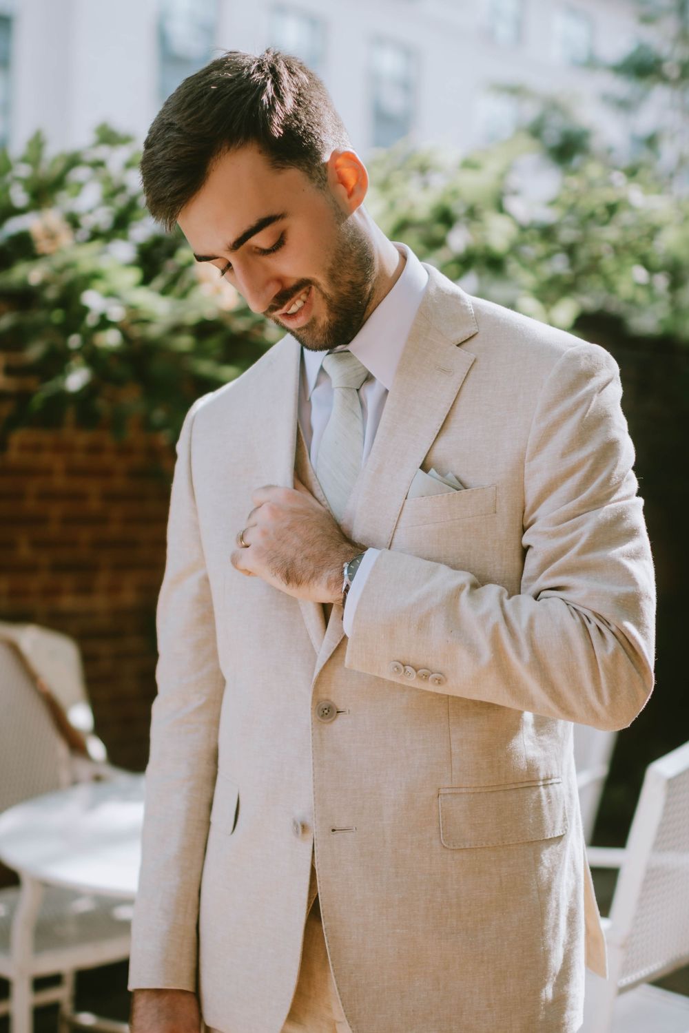 A well-dressed man in a beige linen suit adjusts his jacket outdoors, smiling downward on a sunny day.