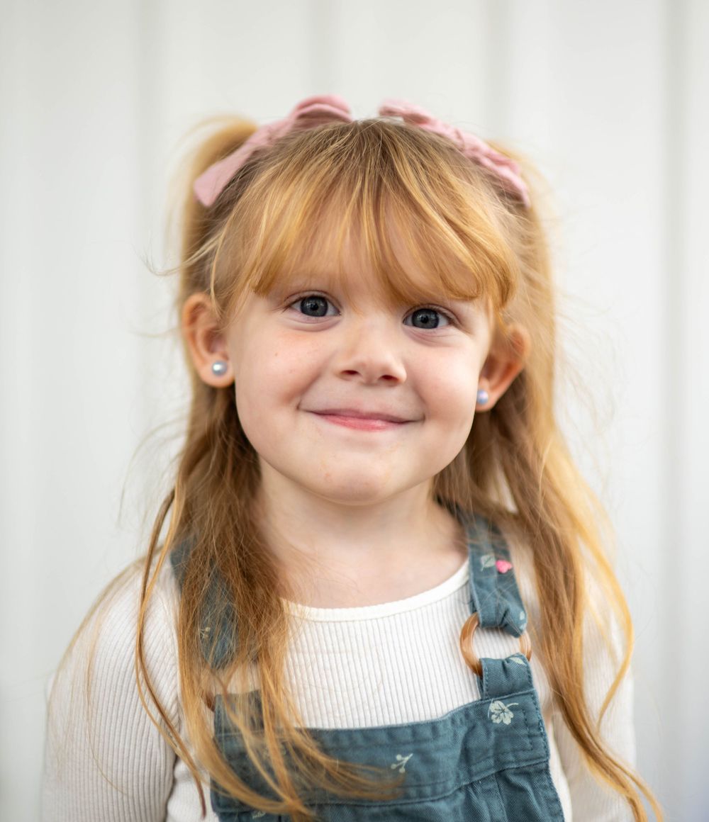 A smiling young girl with red hair in pigtails, pink bows, bangs, wearing a denim jumper over a white shirt.