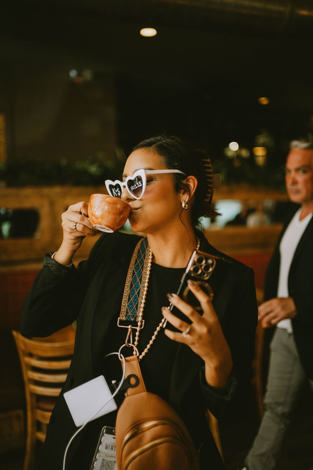 A stylish woman in a black blazer sips from an orange cup while holding a phone, wearing white heart-shaped sunglasses indoors.