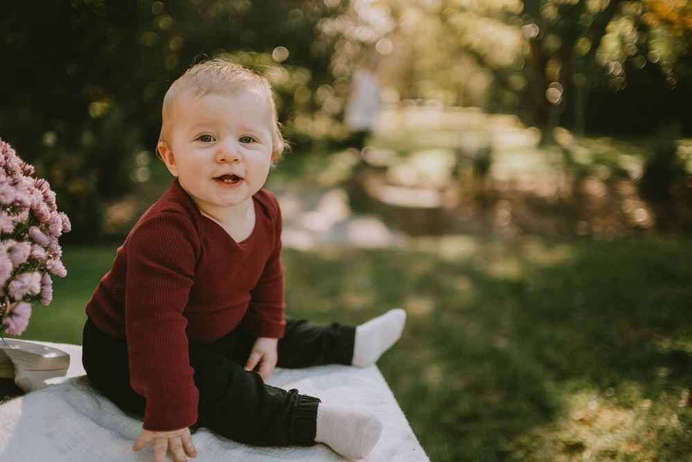 A smiling baby in a red sweater and dark pants sits outdoors on a white surface surrounded by green trees and pink flowers.