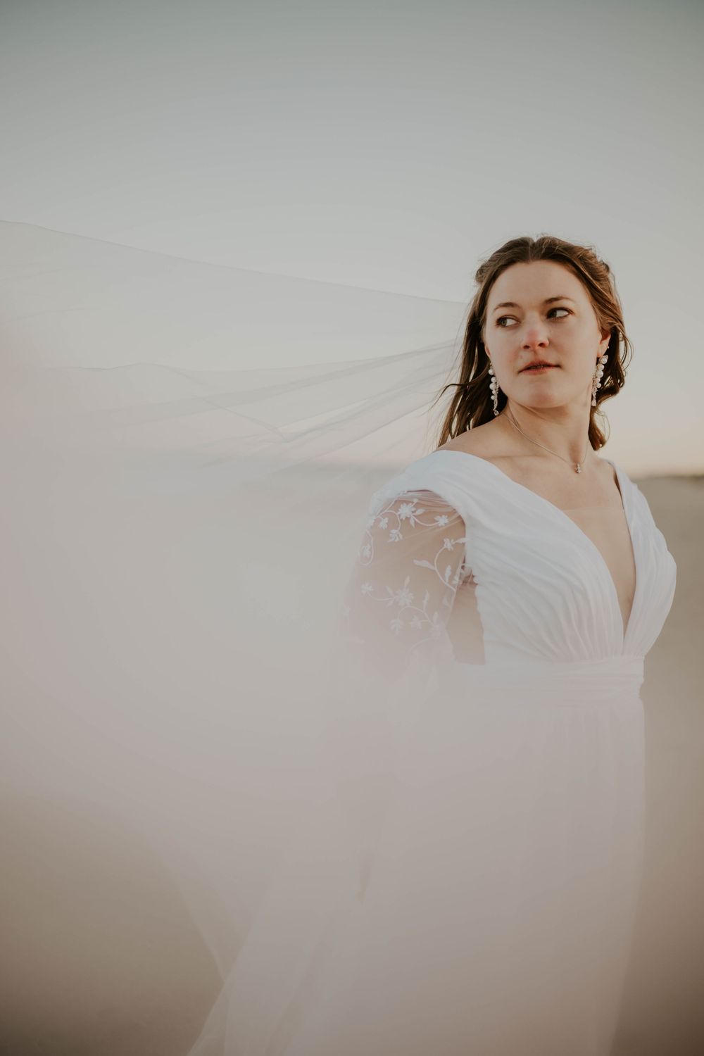 Bride in white lace wedding dress with flowing veil blowing in wind against soft sky backdrop.