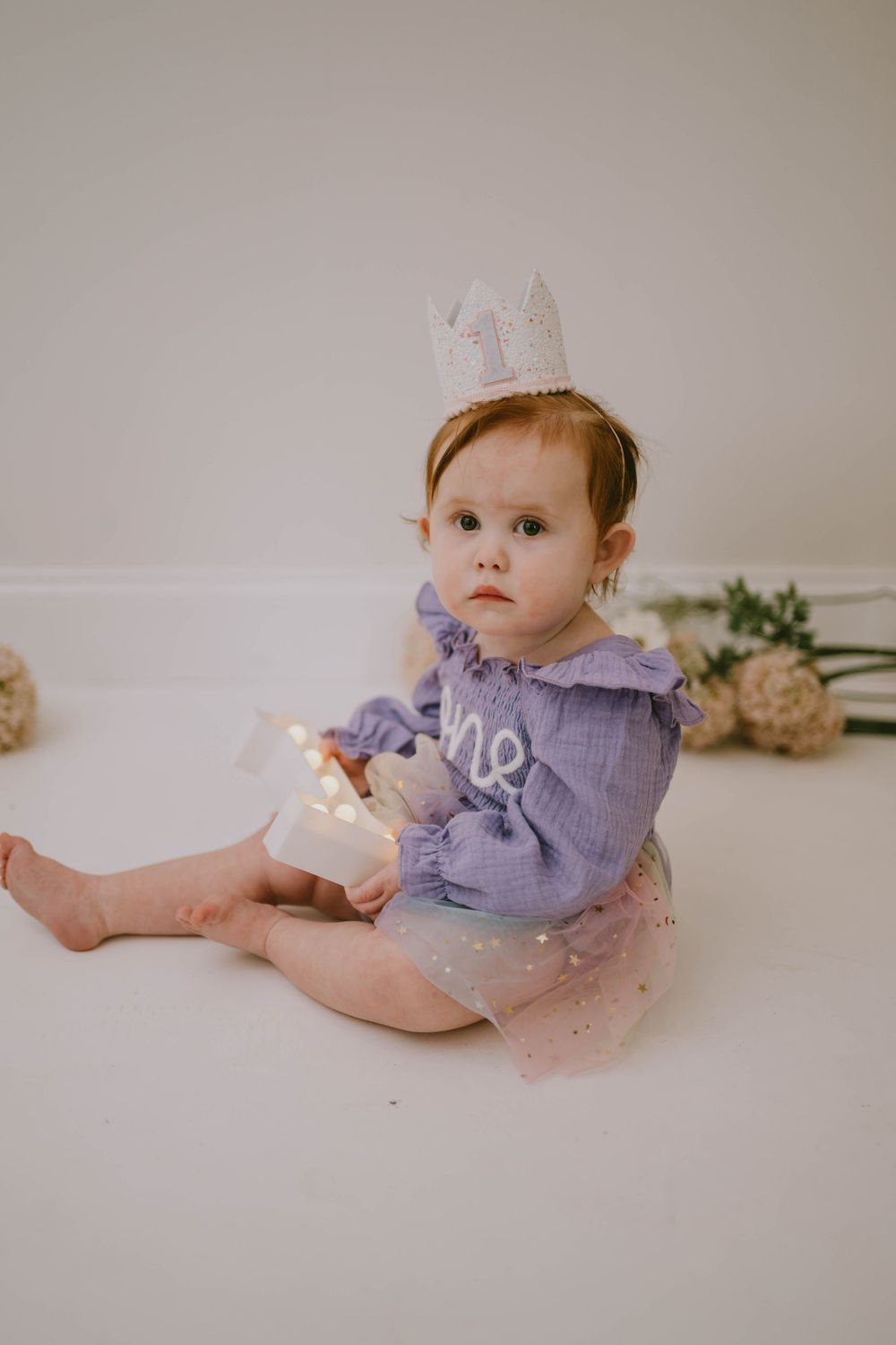Baby girl in purple ruffle romper and glitter crown sits on white floor holding lights at her first birthday.