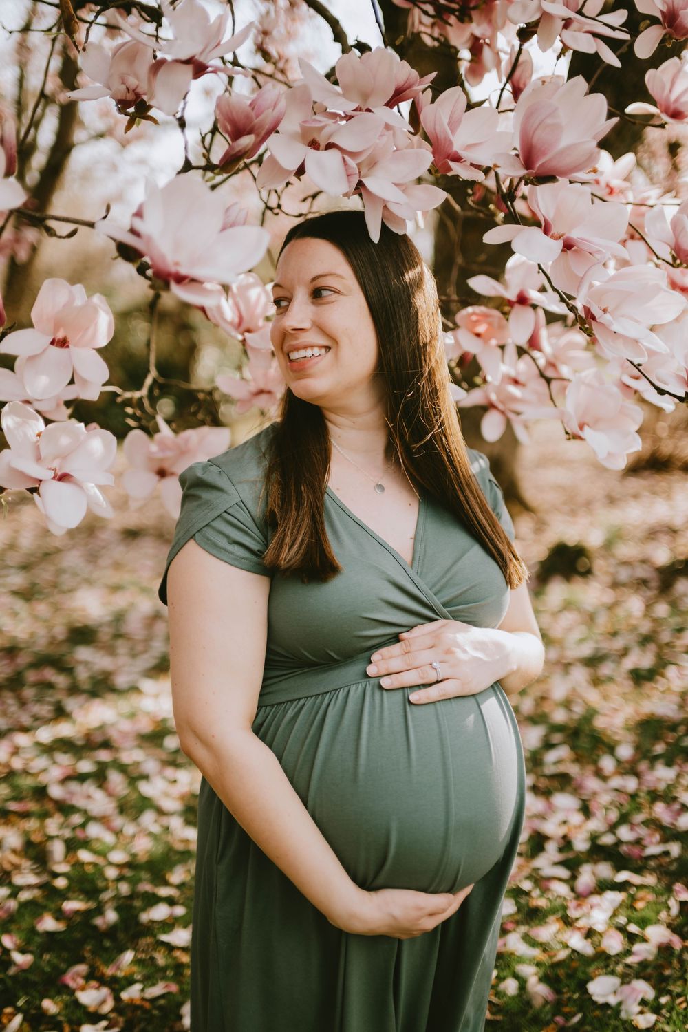 Smiling pregnant woman in green dress cradling baby bump under pink magnolia blossoms in spring.