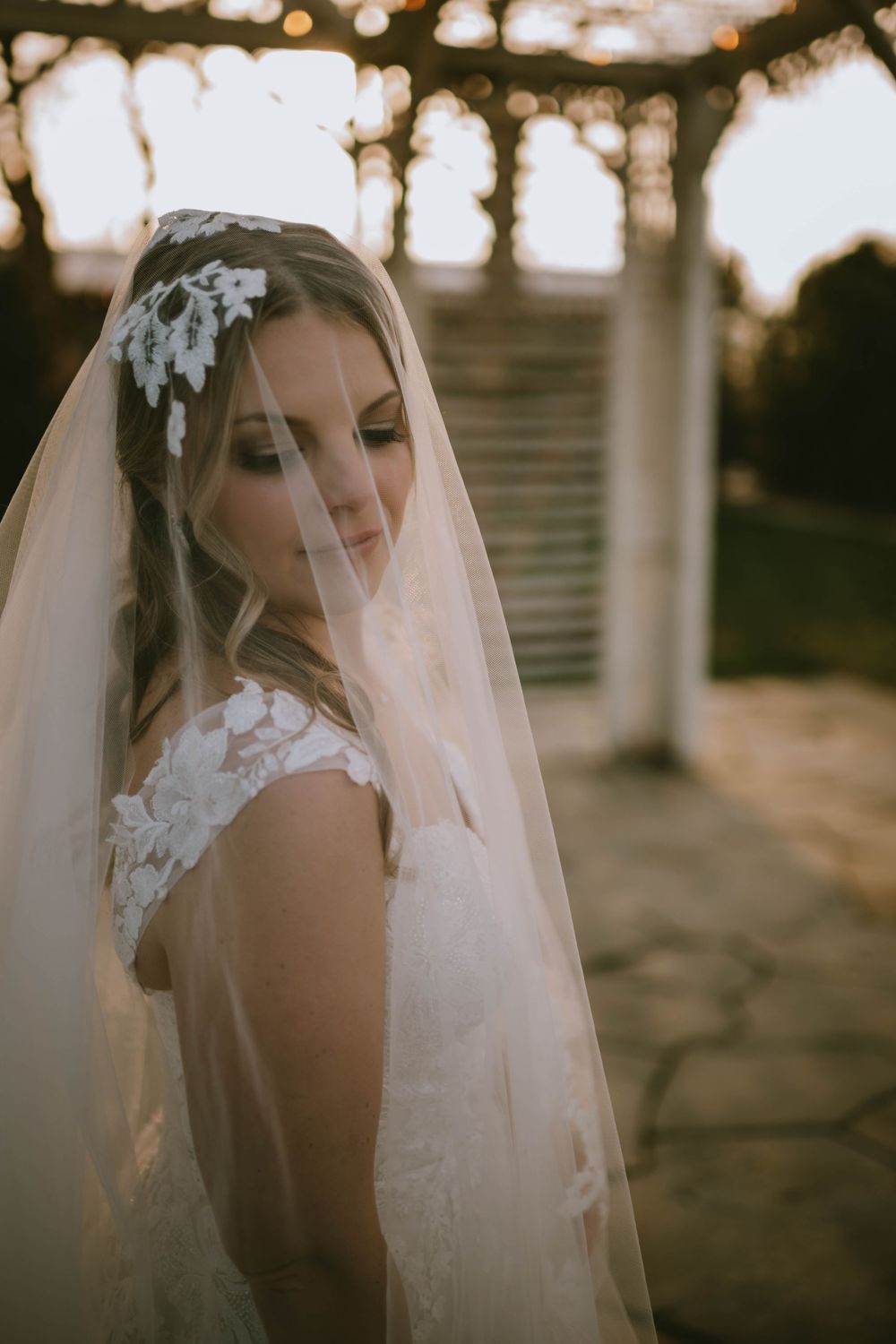 Bride in lace wedding dress and veil with floral hairpiece poses gracefully outdoors at golden hour.