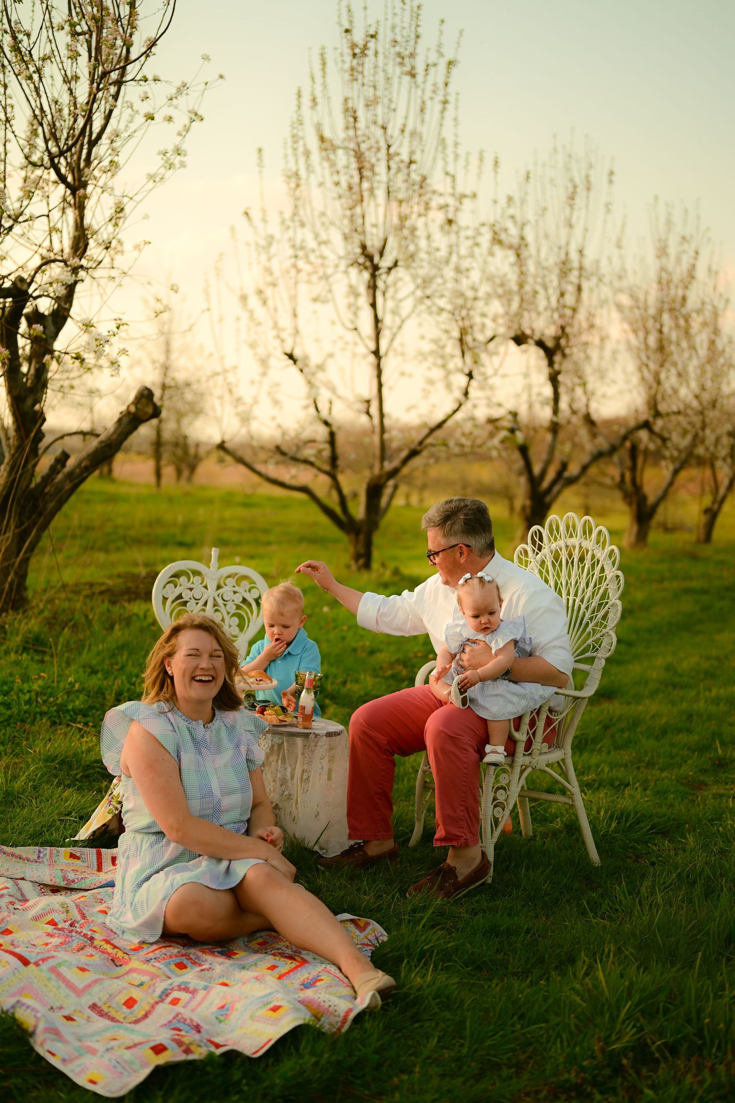 Family enjoying a sunny outdoor picnic in an orchard, with grandparents laughing and holding babies on a colorful blanket.