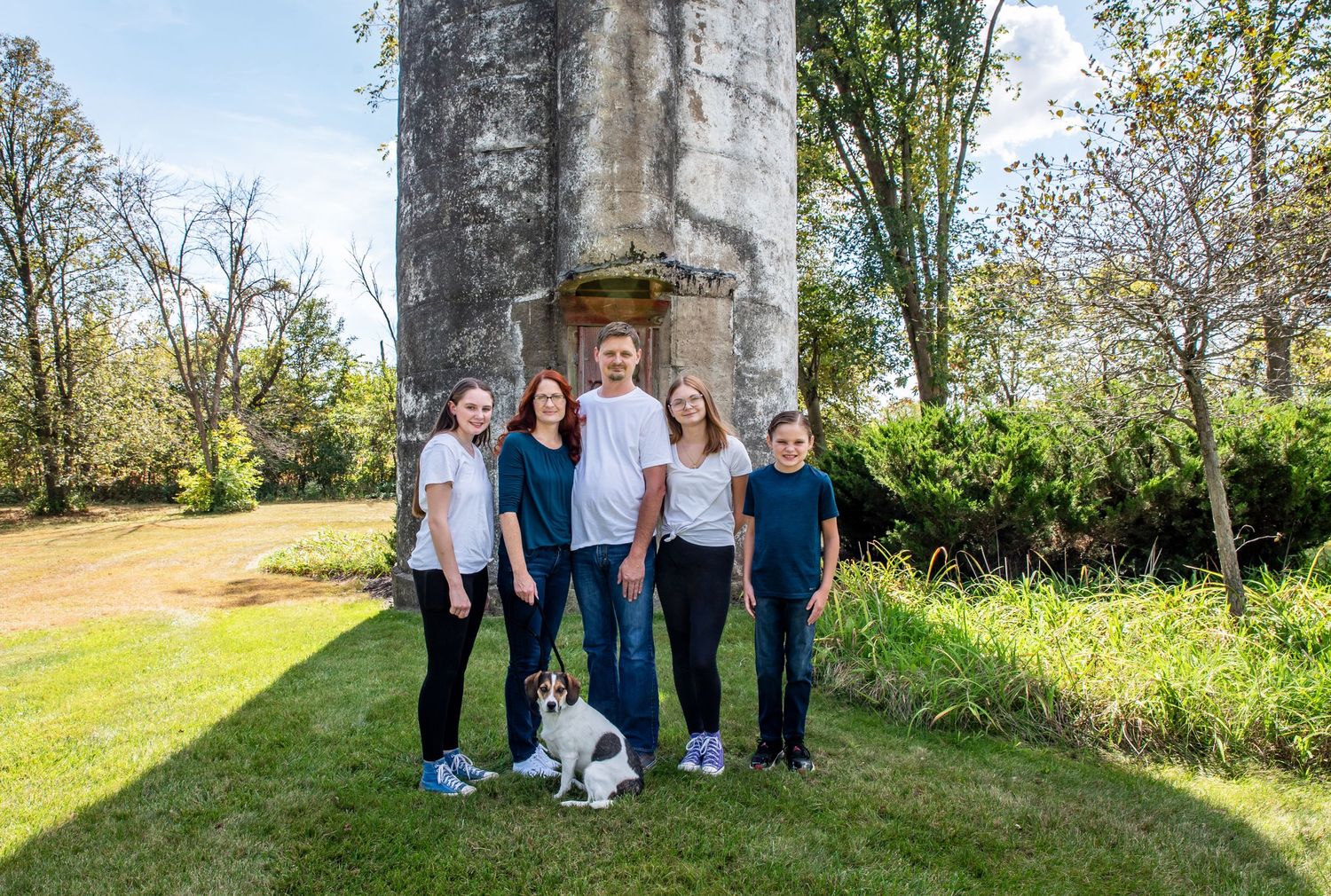 Family group portrait with a small dog in front of a tall concrete structure on a sunny day.