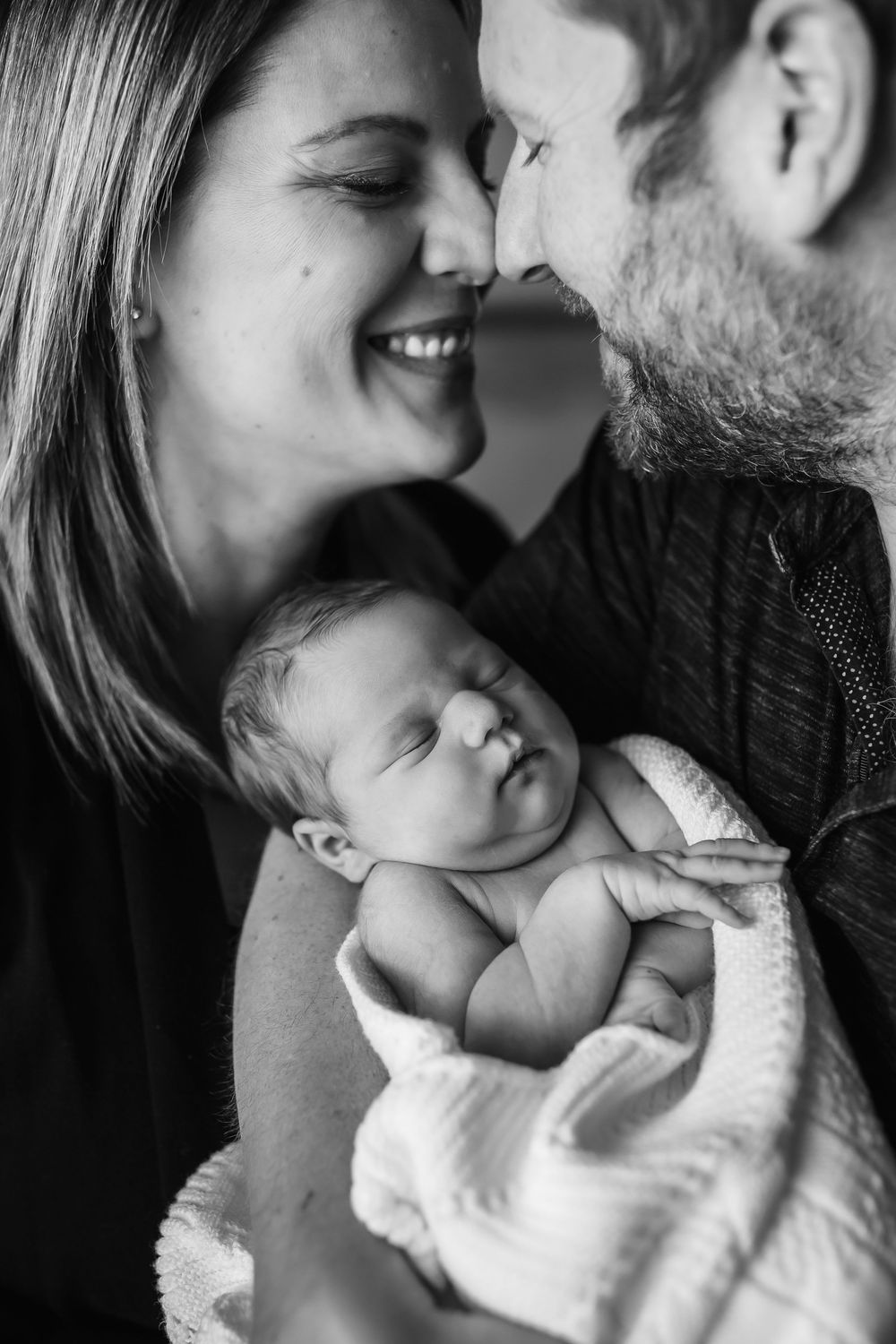 Parents smile joyfully while holding their swaddled newborn in black and white portrait