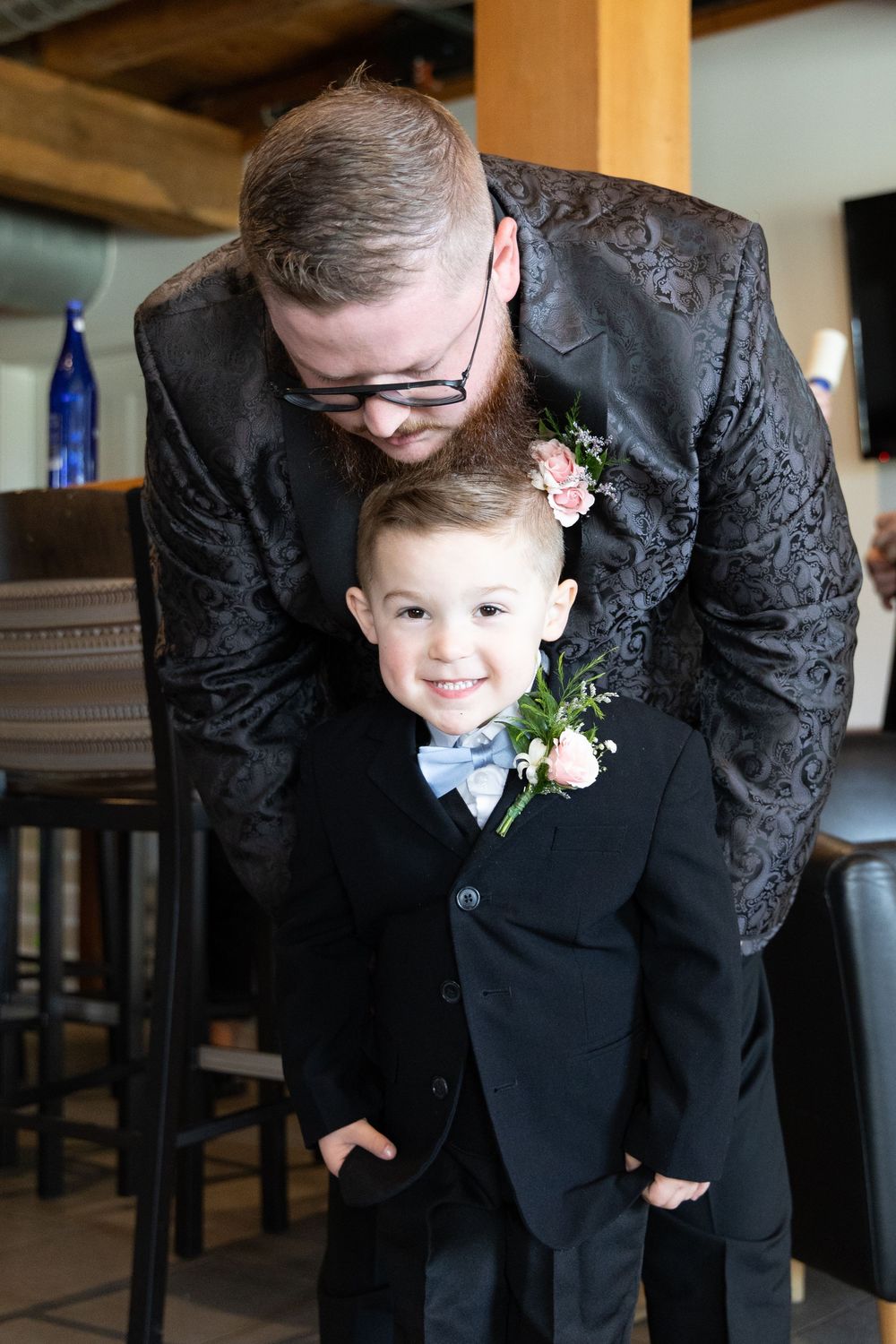 A person in a black patterned suit jacket bends down next to a small child in a formal suit at a special event.