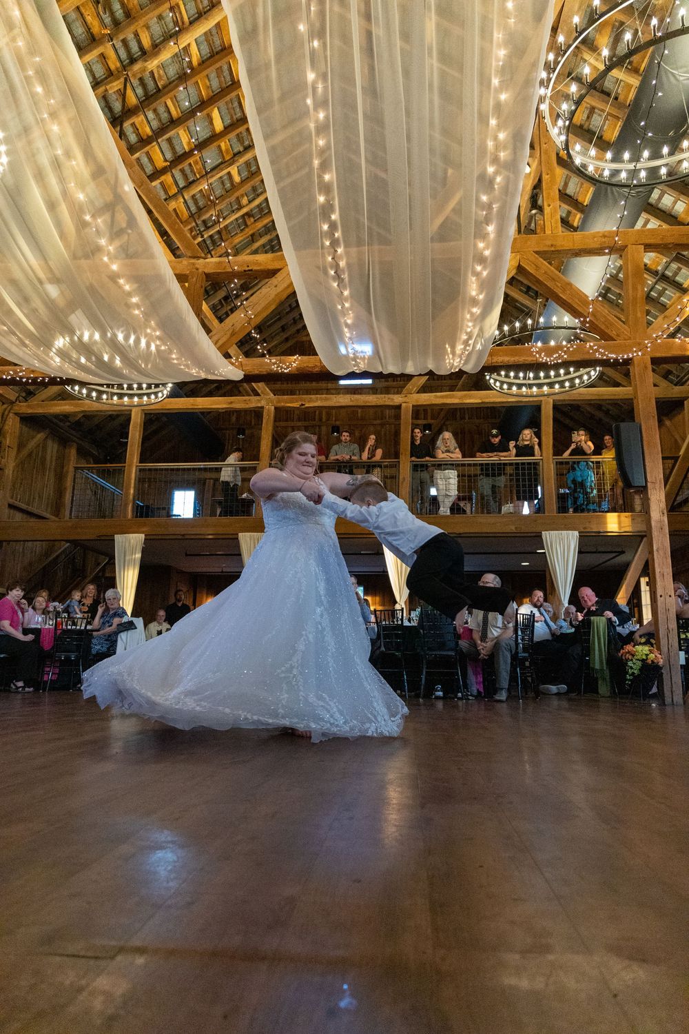A couple dances their first dance under draped fabric and string lights in a rustic barn venue.