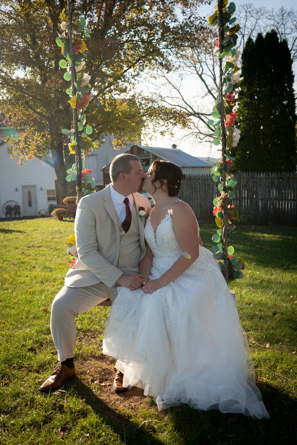 A romantic outdoor wedding scene with a couple embracing on a wooden bench surrounded by fall trees and string lights at sunset.