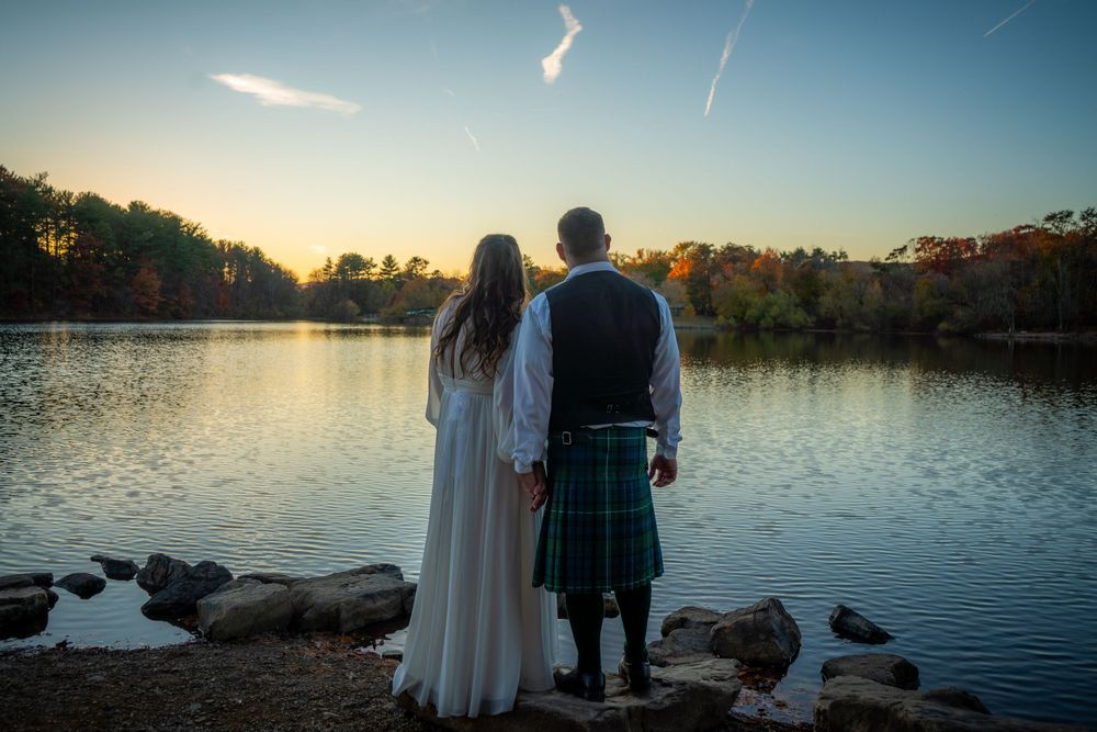 A couple in traditional Scottish attire stands at the edge of a tranquil lake during sunset.
