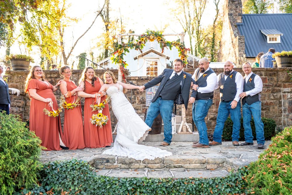 A wedding party poses outdoors in a line against a stone wall with bridesmaids in coral dresses and groomsmen in blue.