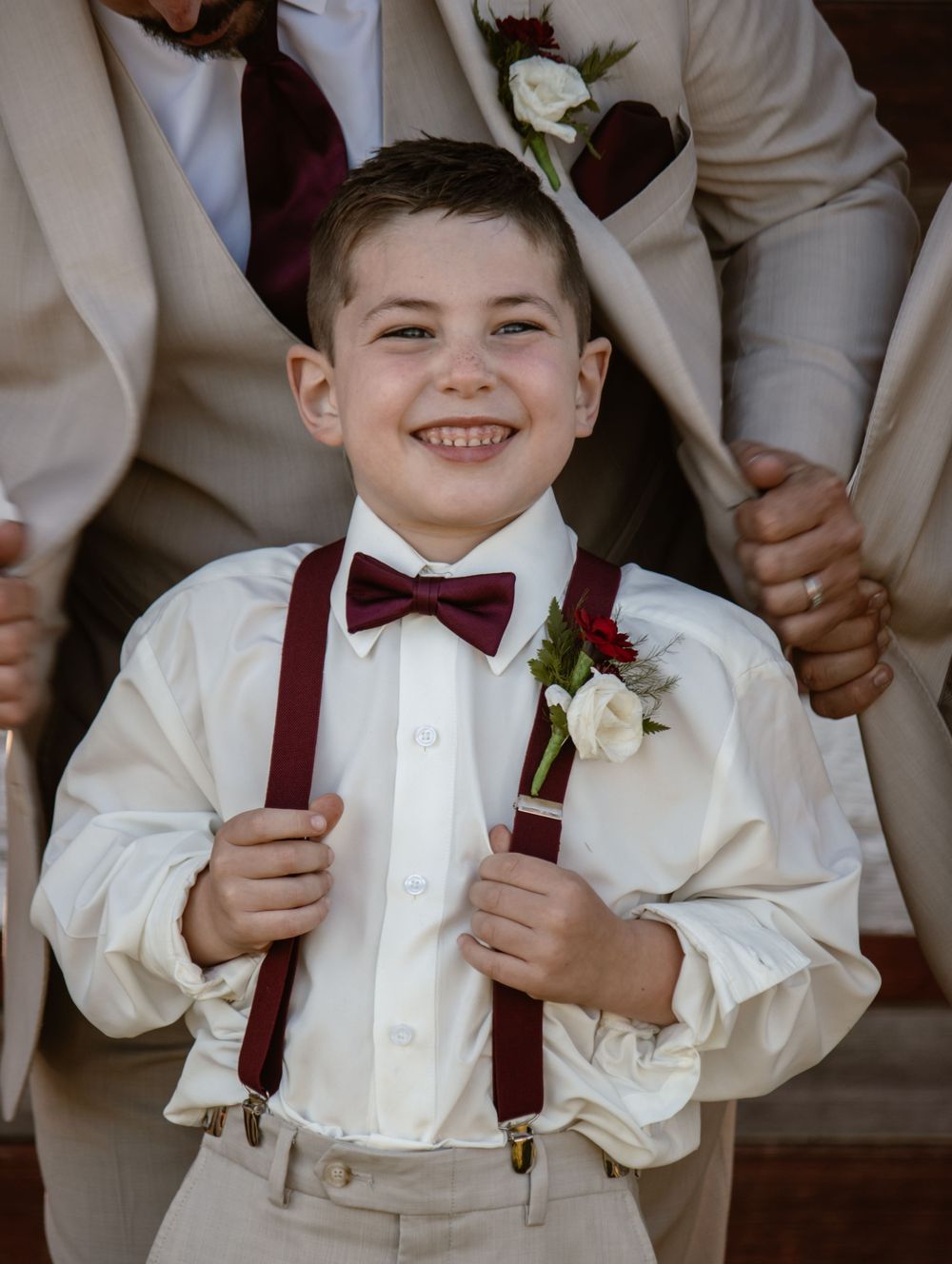 A young ring bearer in white shirt and burgundy suspenders smiles while holding a boutonniere at a wedding.