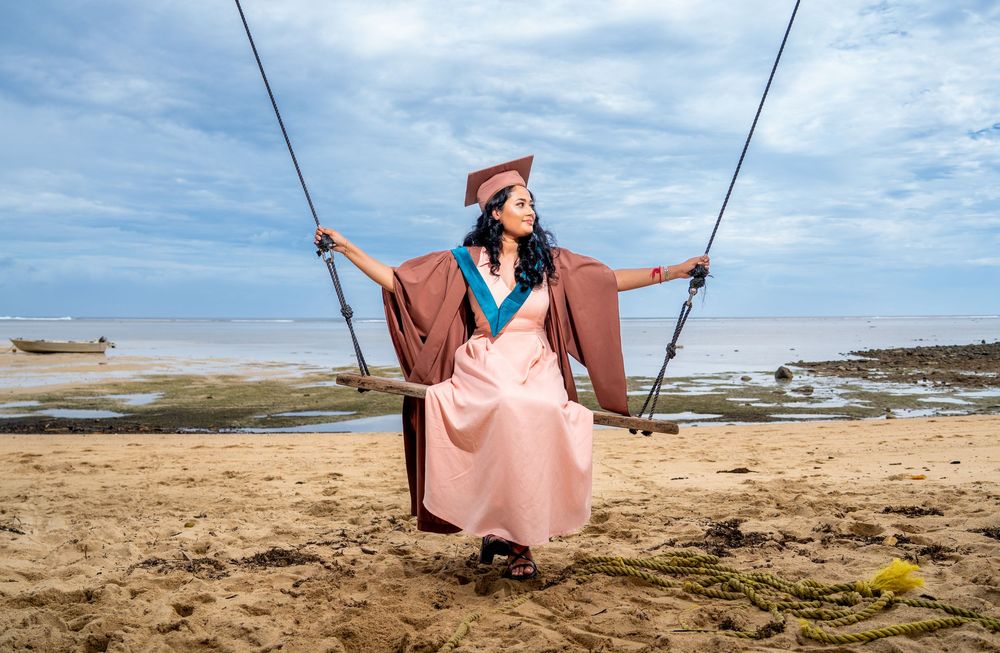 A person in a flowing brown coat and pink dress sits on a swing against a coastal landscape with blue skies.