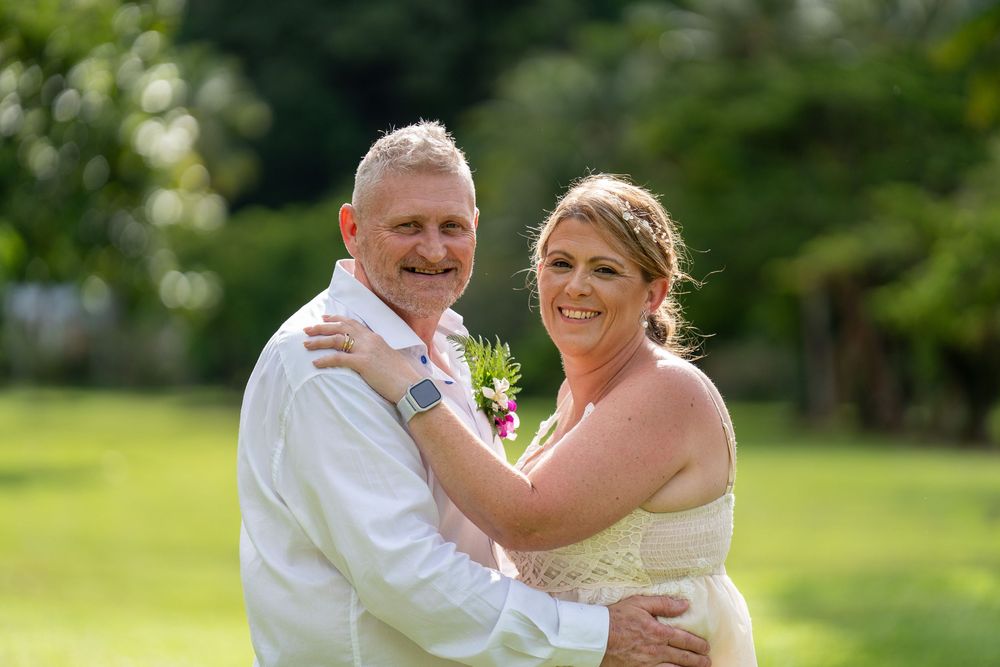 A happy couple embraces and smiles while celebrating their wedding day in a sun-lit outdoor garden setting.