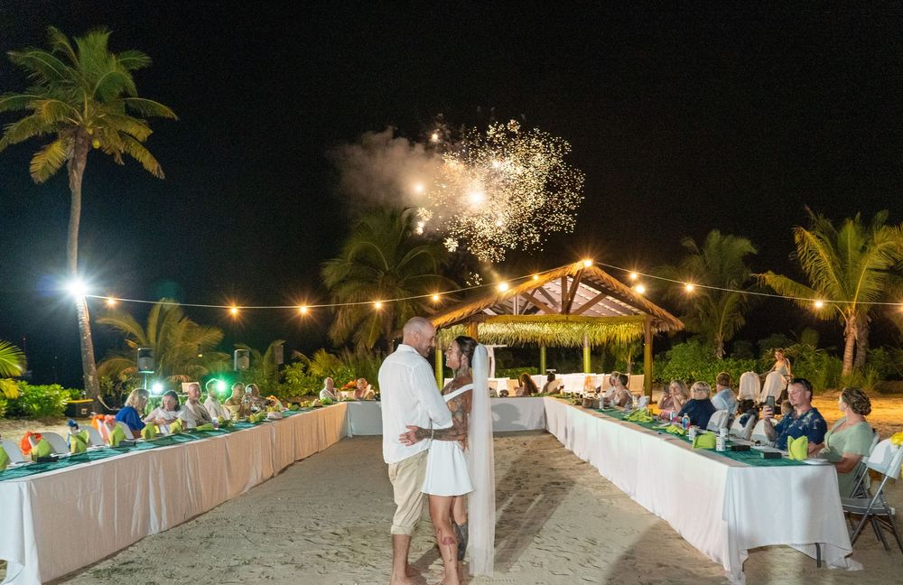 Newlyweds watch fireworks display during nighttime beach reception with palm trees and lit tables on the sand.