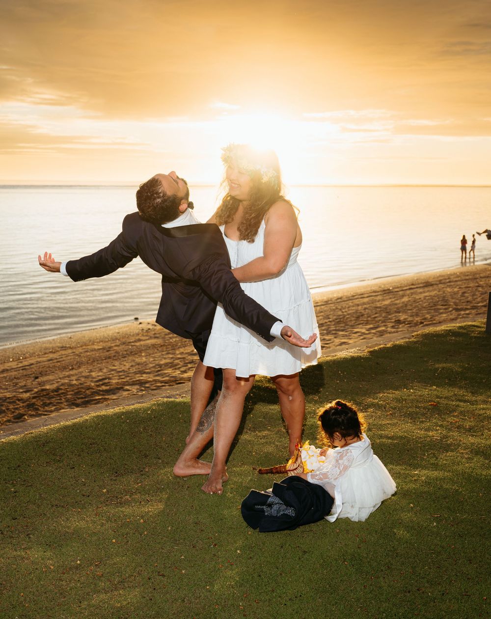 A couple dances together on a grassy hill overlooking a beach at sunset while their belongings rest nearby.