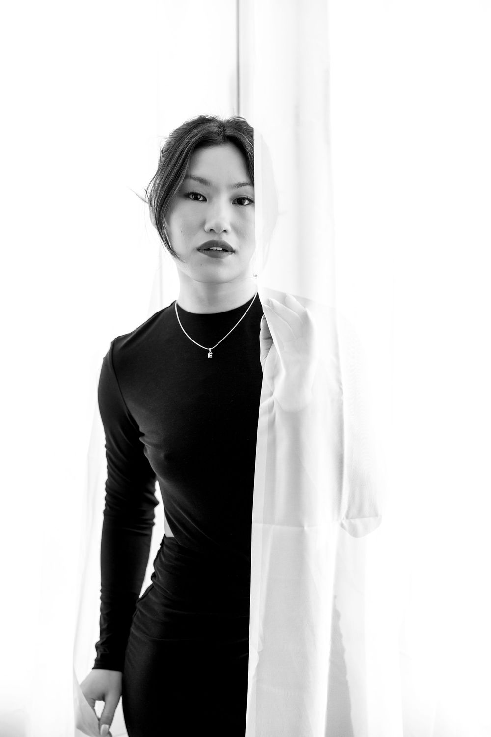 Black and white portrait of Asian woman in black dress holding white sheer curtain by window.