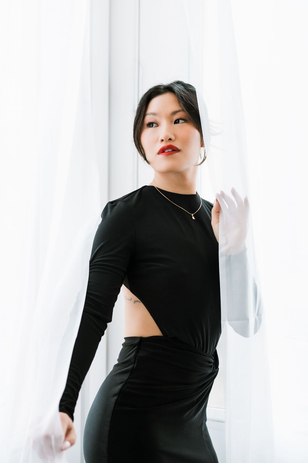Asian woman in black cutout dress posing elegantly beside white sheer curtains in bright room.