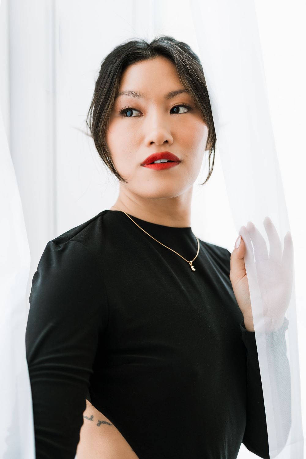 Asian woman with red lips in black long-sleeve dress holding white curtain, small tattoo visible.