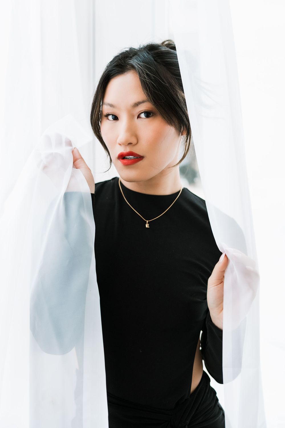 Asian model in black fitted dress parting white sheer curtains, wearing gold pendant necklace.
