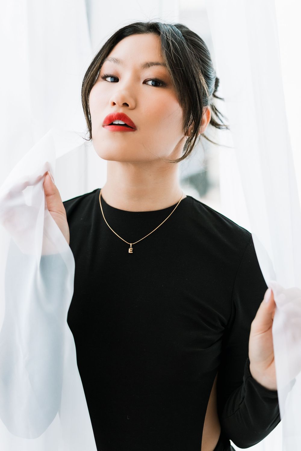 Asian woman in black dress with cutout detail holding white curtains, gazing upward gracefully.