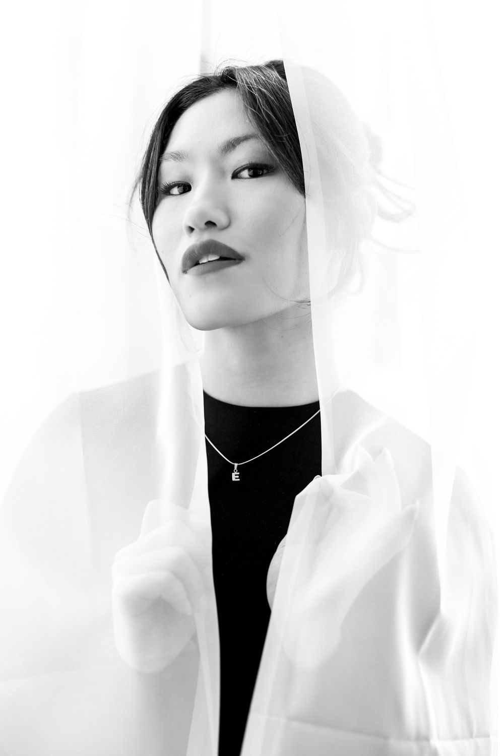 Asian woman in black long-sleeve dress wrapped in white sheer fabric, wearing gold necklace.