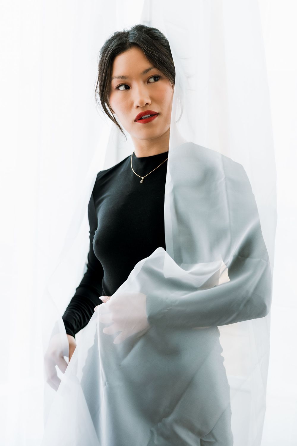 Black and white close-up portrait of Asian woman with sheer white curtain, delicate necklace.