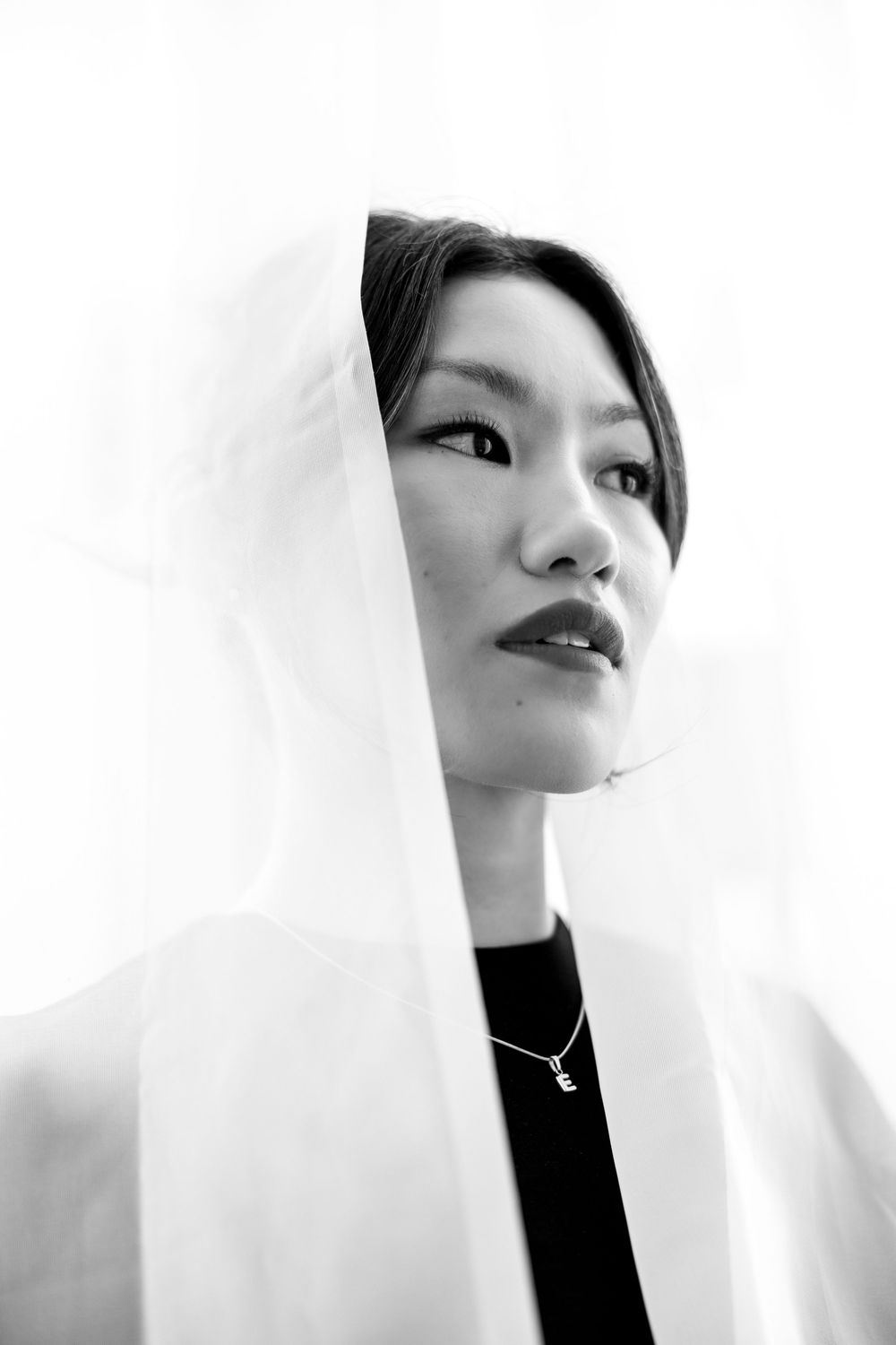 Asian woman in backless black long-sleeve top looking over shoulder, red lips, white background.