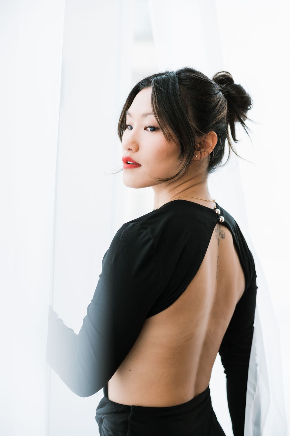 Young Asian woman in backless black top with red lips and hair up, posing elegantly against white curtains.