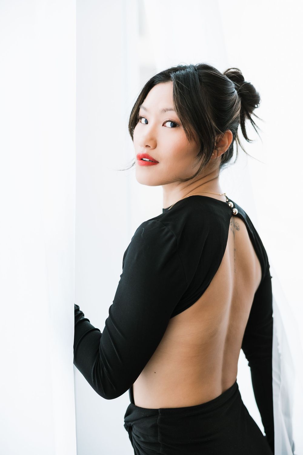 Woman in backless black dress posing elegantly with red lips and updo hairstyle against white background.