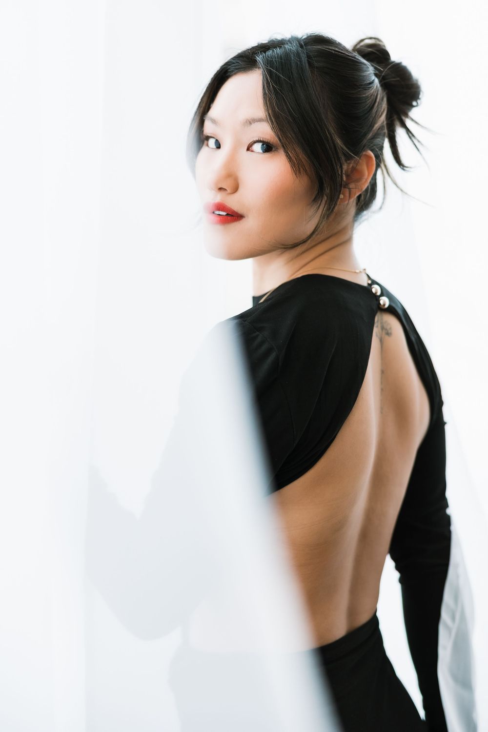 Elegant Asian woman in open-back black dress posing gracefully near white curtains in soft light.