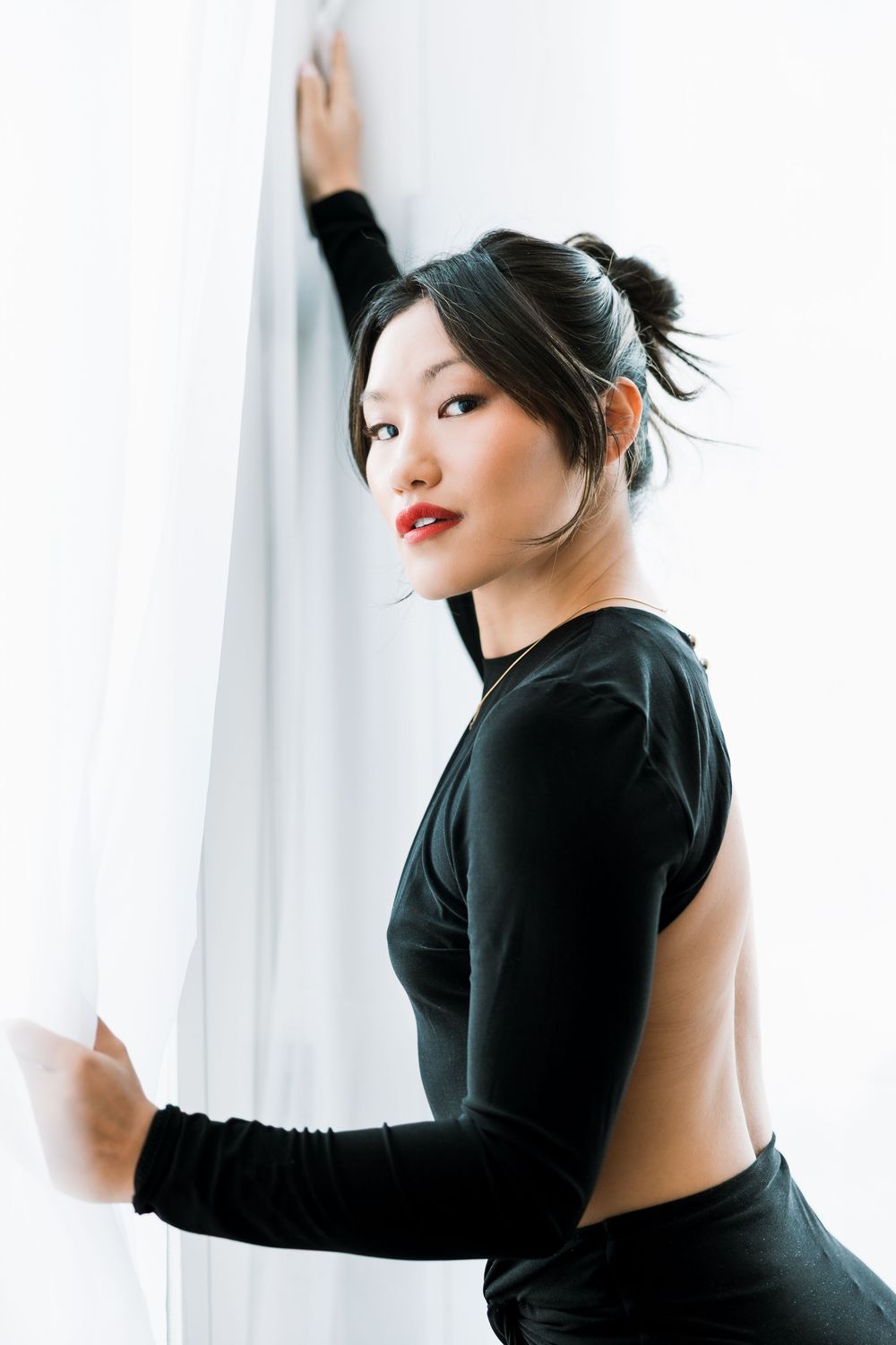 Stylish woman in backless black outfit reaching up to hold white curtain in bright studio setting.