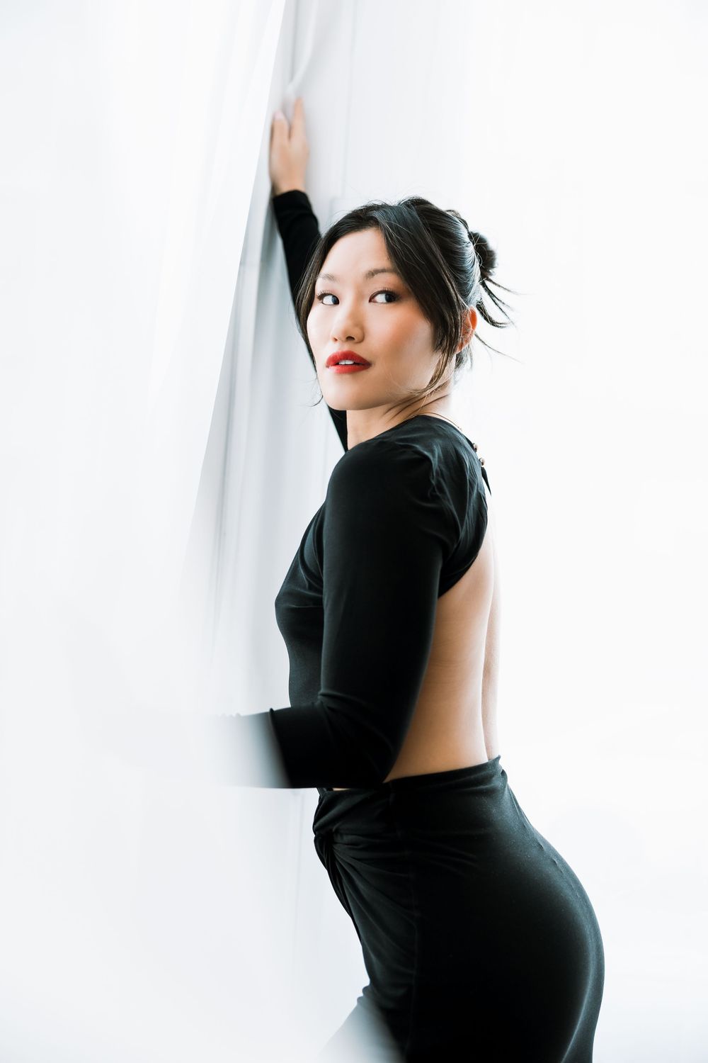 Fashion model in backless black dress posing beside white curtain with red lips and sleek updo.