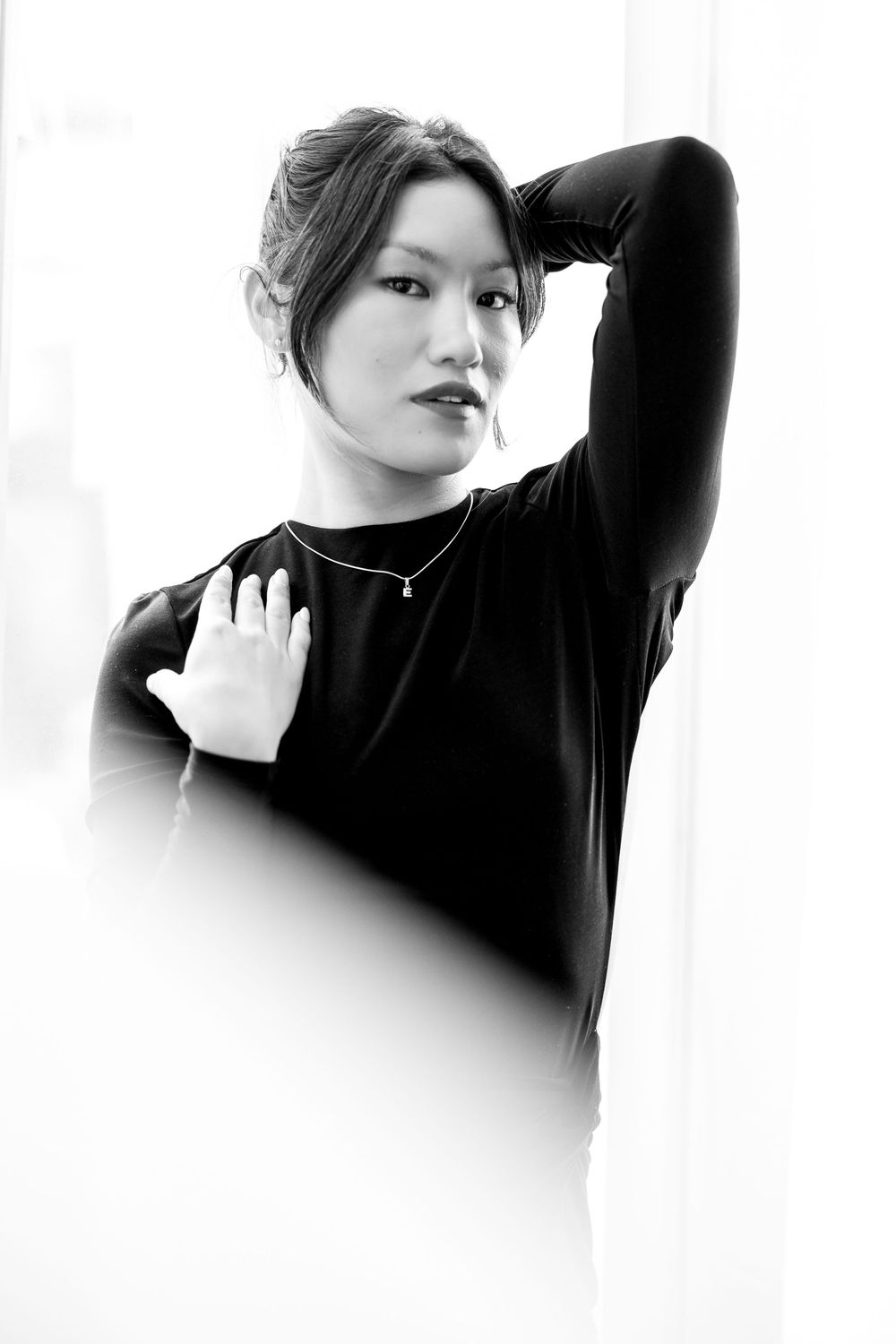 Black and white portrait of woman in black top with necklace posing near bright window.