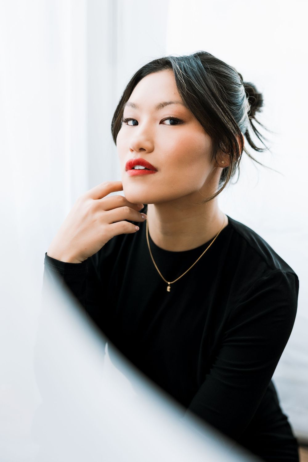 Close-up portrait of Asian woman with red lips and gold necklace gazing upward near white wall.