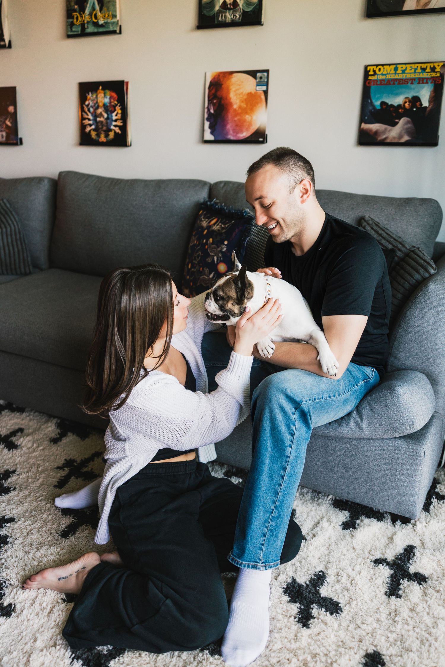 Smiling couple sits on a gray sofa petting a small French Bulldog in a cozy living room.