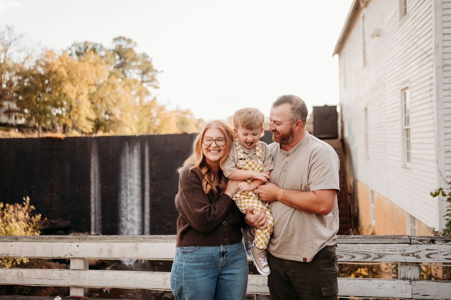 Happy family of three smiling outdoors in autumn, parents holding toddler near rustic white building.