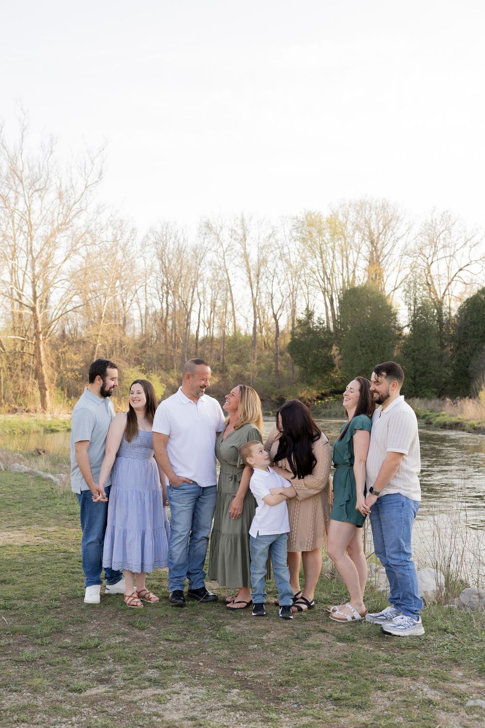 A group of people gather for photos on a grassy bank beside a river at river bends park in utica during a casual outdoor photoshoot.