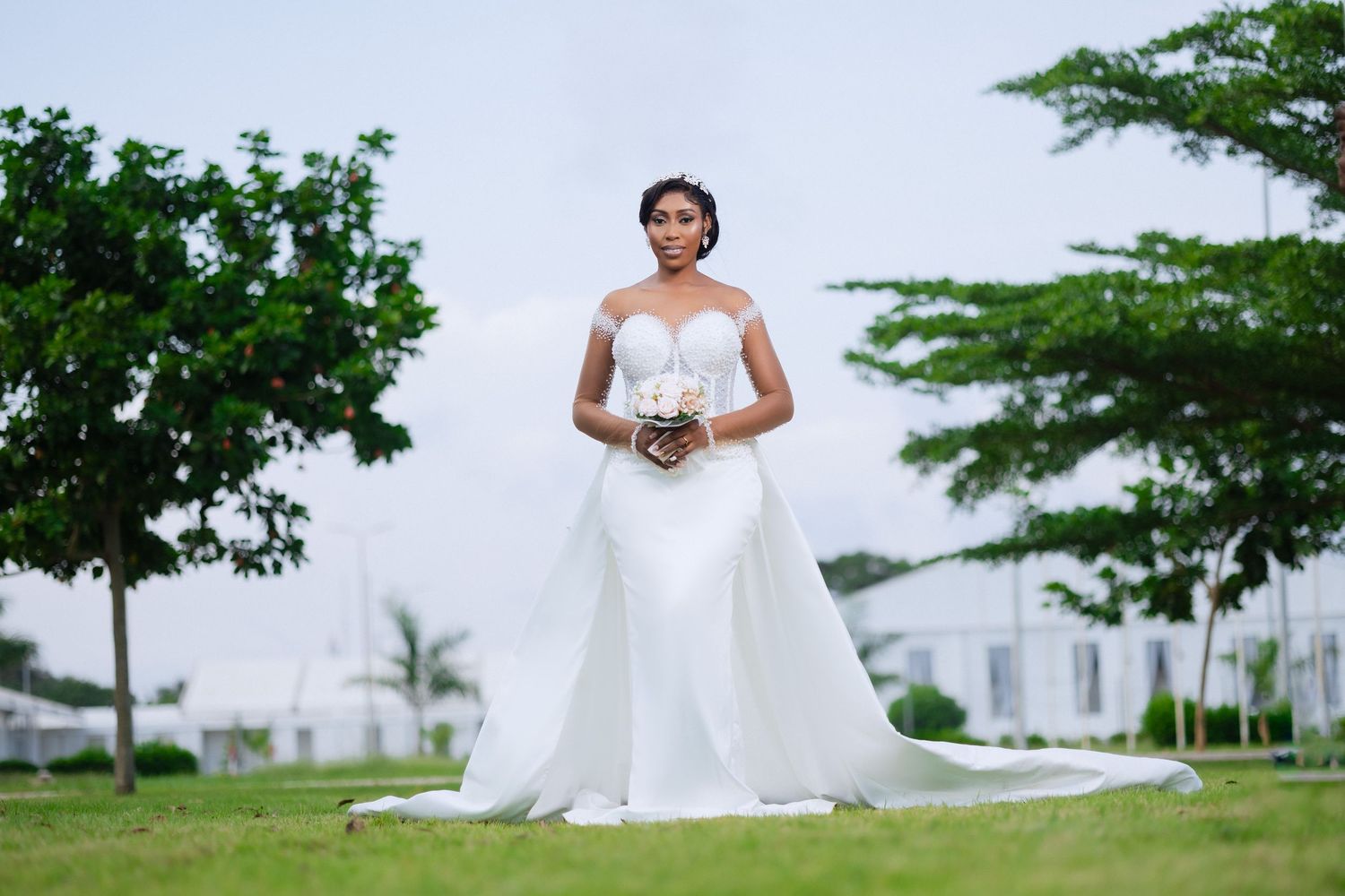 A bride in a white wedding gown poses in a lush garden setting with tropical trees and greenery.