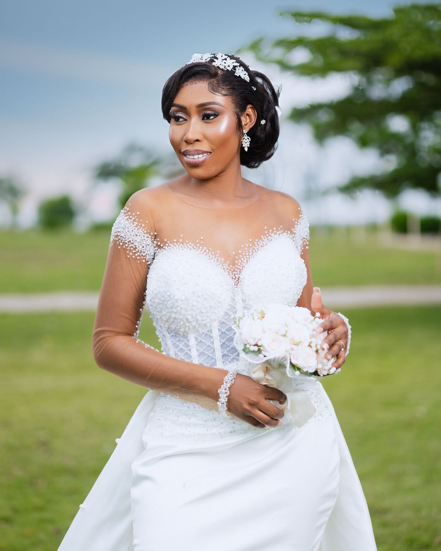 Beautiful bride in off-shoulder white wedding gown with crystal tiara holds white bouquet outdoors on sunny day.