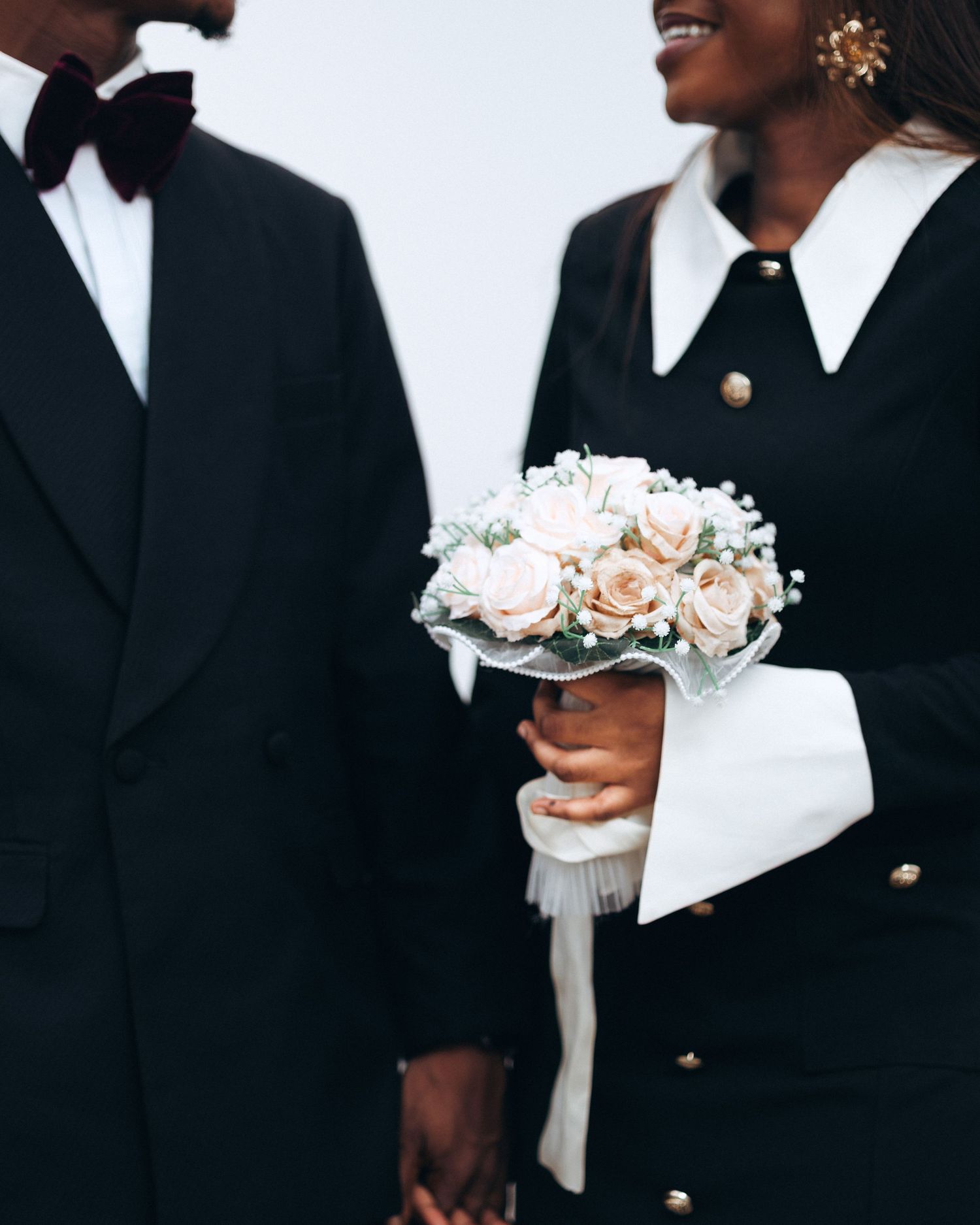 Hands holding delicate pink and white floral bouquets against black formal attire at an elegant wedding ceremony.