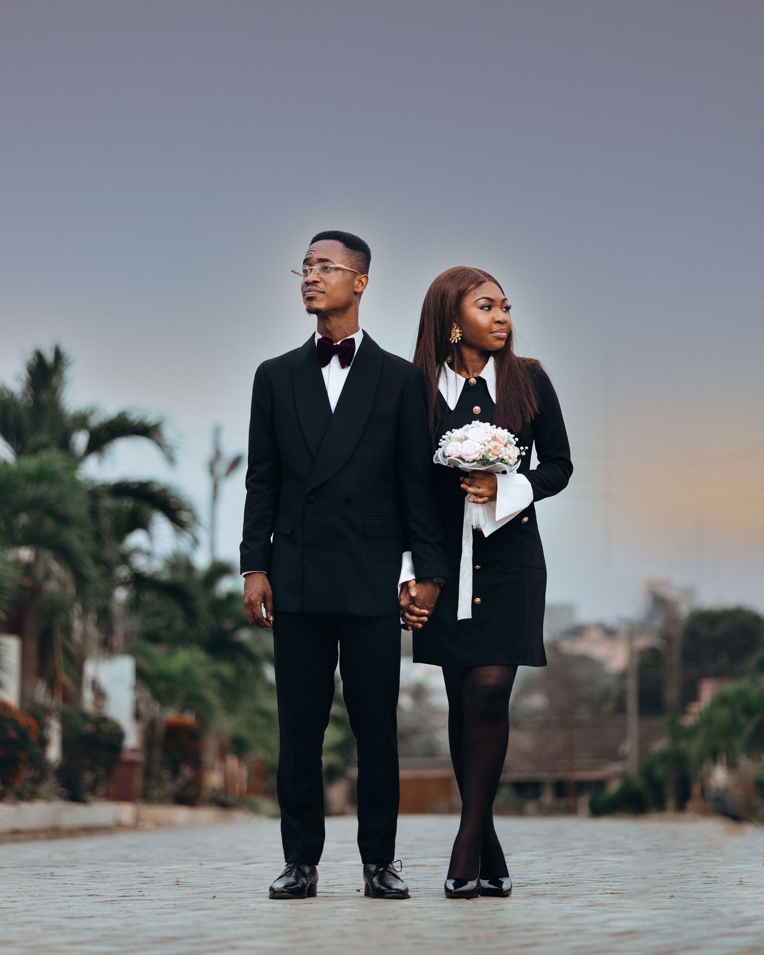 Two pairs of formal shoes stand side by side on a concrete surface with palm trees in the background at sunset.
