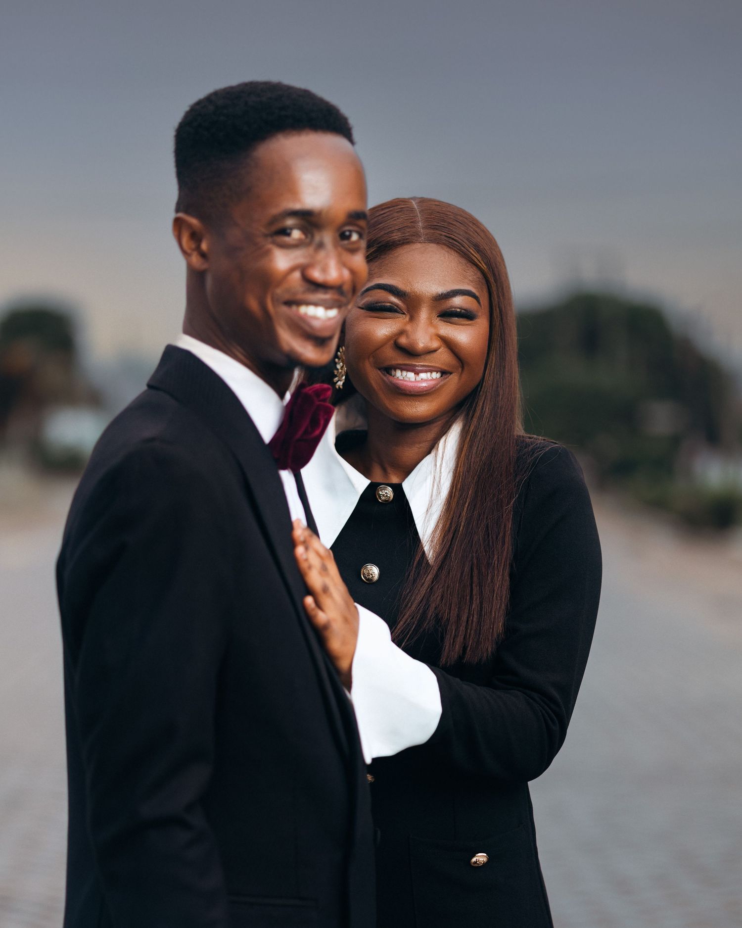 A happy couple in formal attire embrace and smile while posing together outdoors against a blurred background.