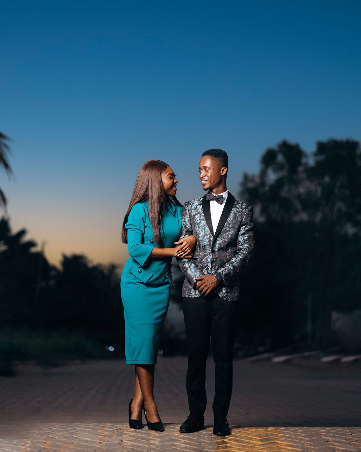 A couple in formal attire share a romantic moment at sunset, with the person in teal dress and other in silver patterned suit.