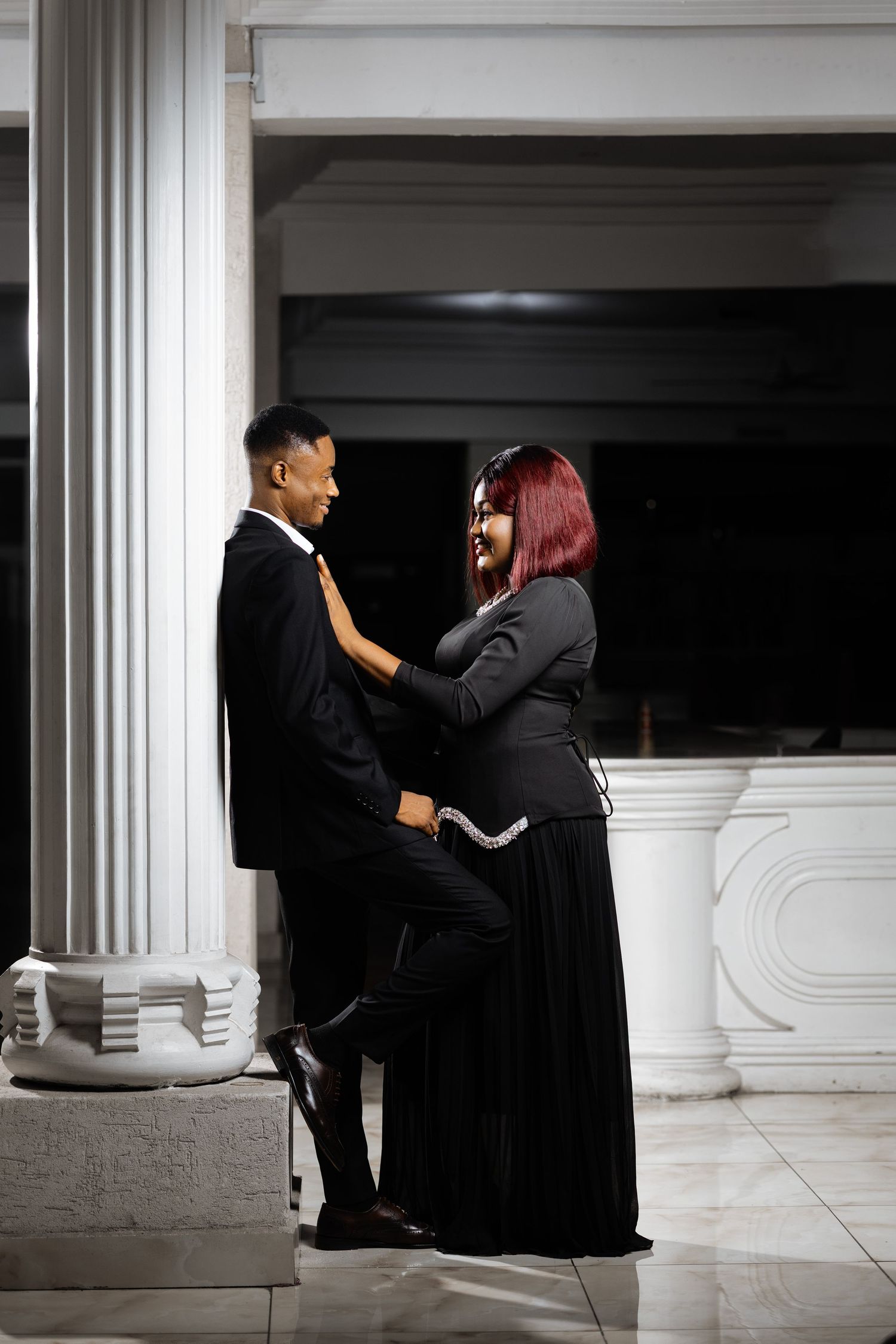 Couple in formal black attire shares an intimate moment against a white pillar.