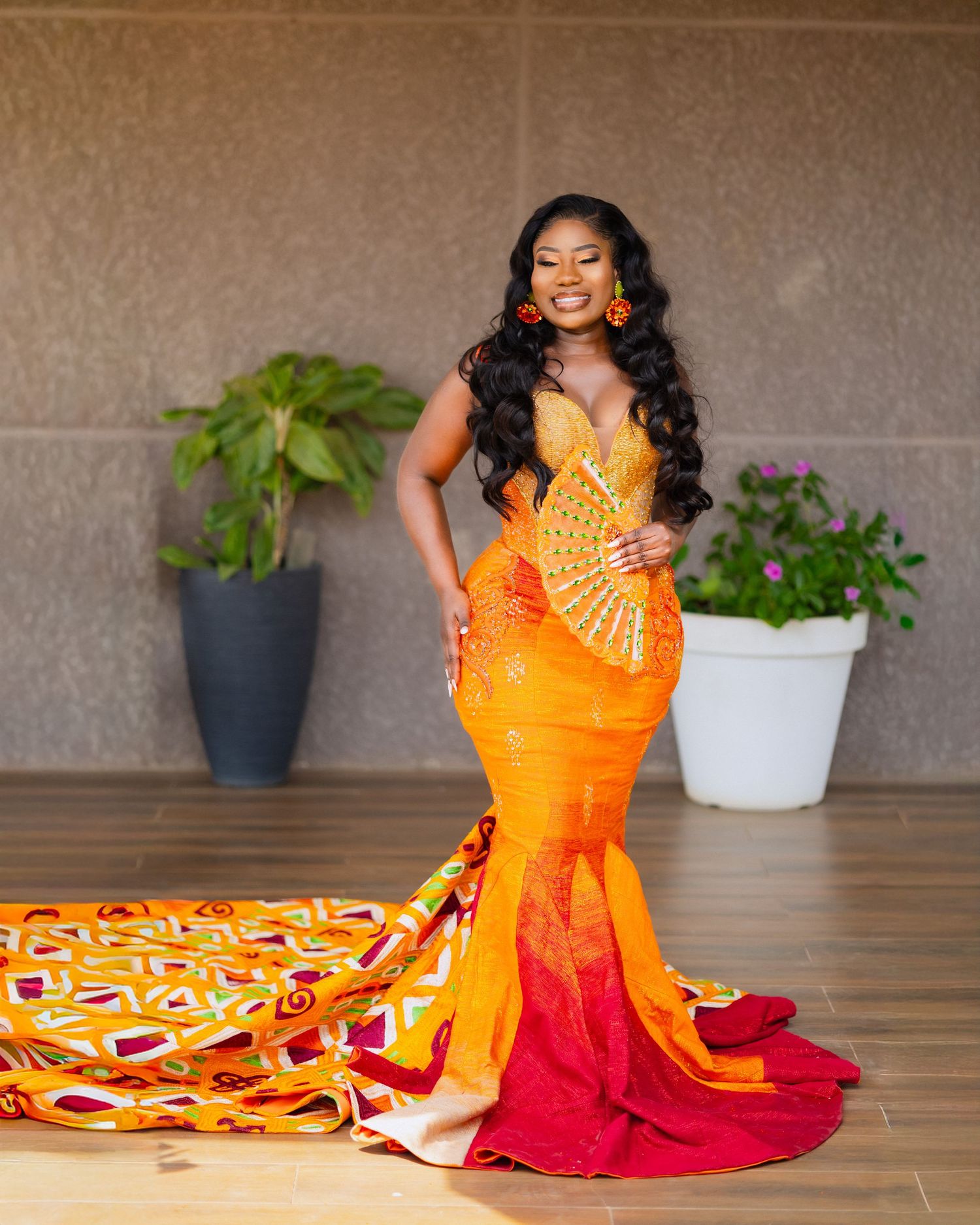 A figure in a vibrant orange and red traditional dress poses gracefully on a floor with potted plants in the background.