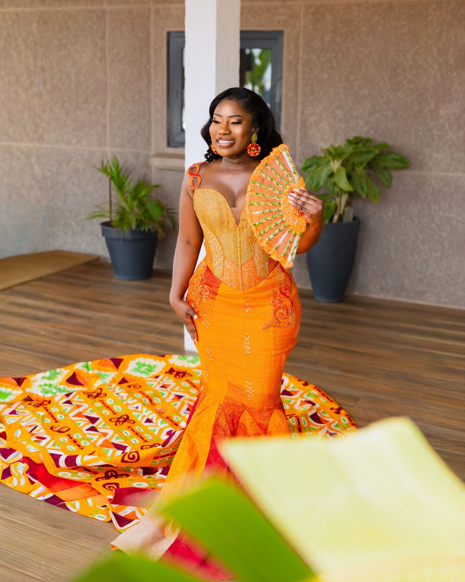 Person in vibrant orange dress with floral details poses on wooden floor near potted plants.